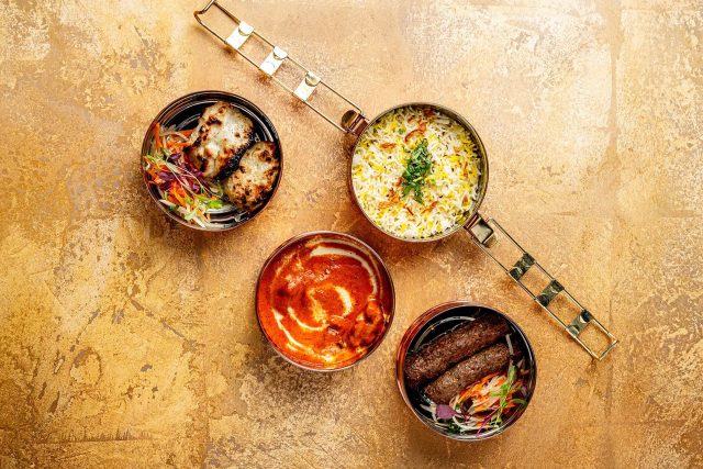Overhead view of an open copper dinner dabba displaying a selection of Indian dishes including rice, a creamy curry, and kebabs from Masalchi's Chef's Dinner Dabba menu on a textured gold surface.